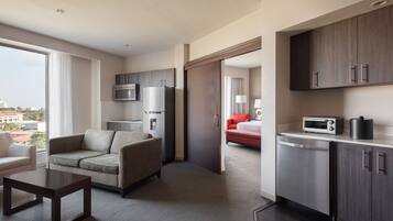 A modern hotel room with a sofa, a small table, a kitchen area with a microwave, refrigerator, and a dishwasher.