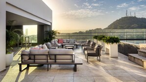 Una terraza en la azotea con mobiliario exterior, plantas en macetas y vistas a una colina con edificios en la distancia.