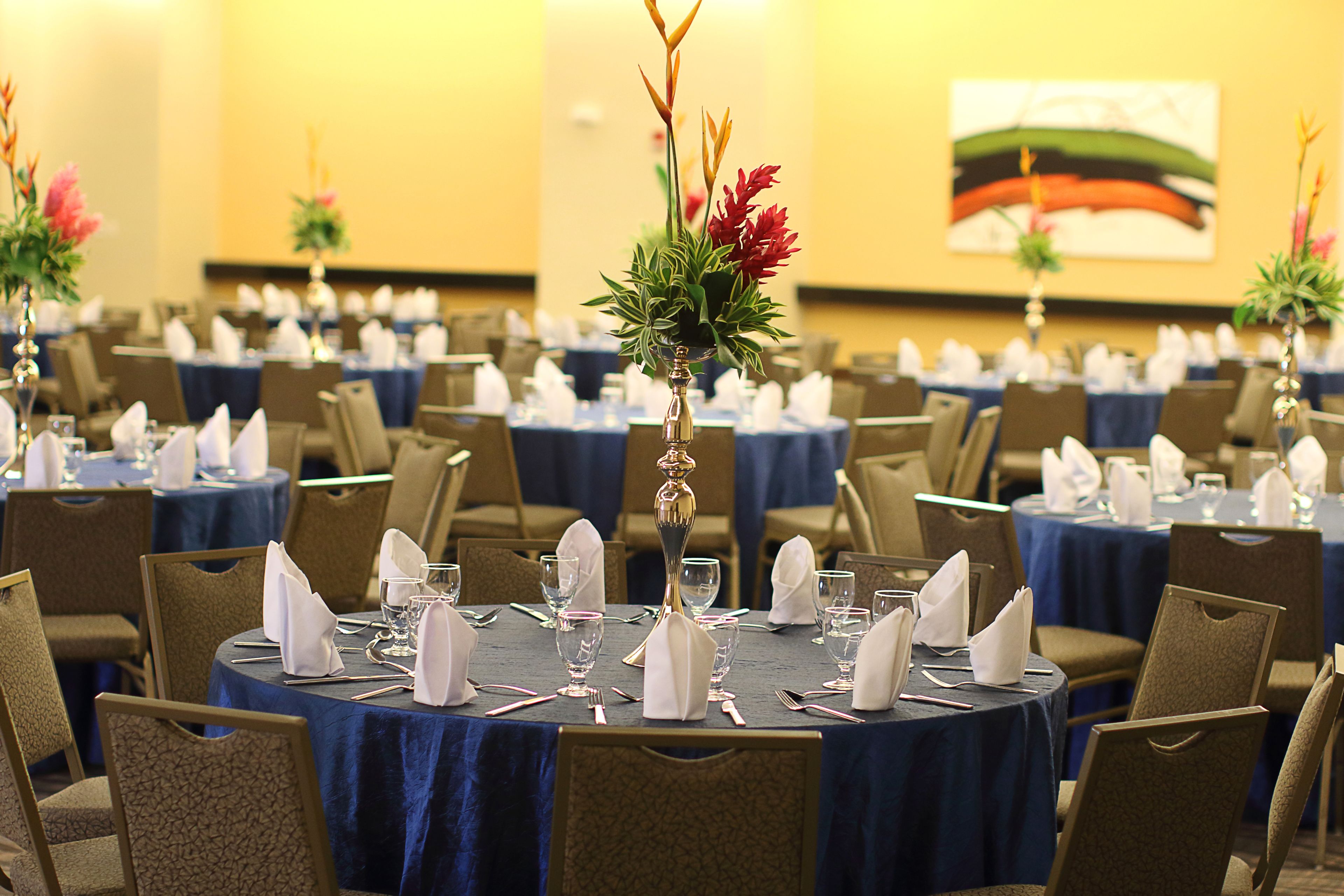Un salón de banquetes con mesas redondas dispuestas para comer, cada una con mantel azul y servilletas blancas.