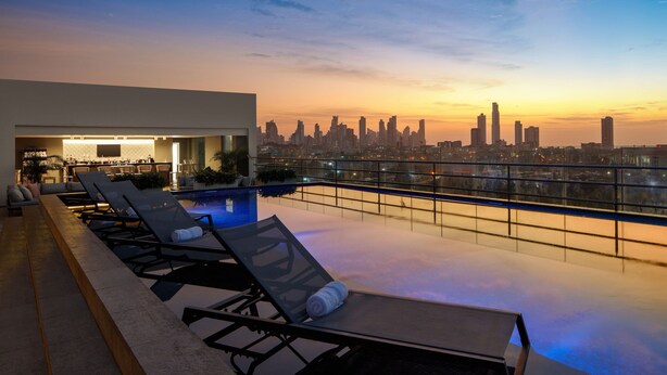 Zona de piscina en la azotea con sillones de descanso y un bar, con vistas a un horizonte urbano al atardecer.