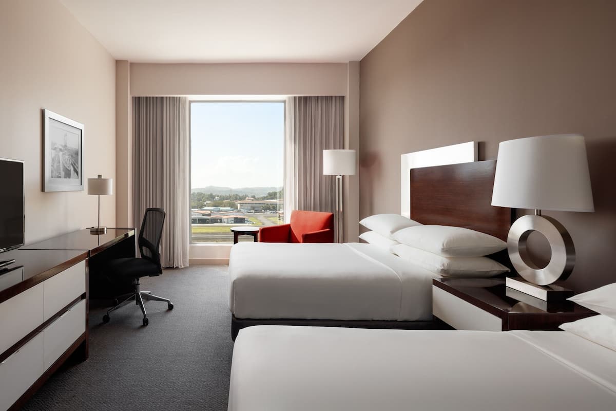 A hotel room with two beds, a desk, a chair, and a TV.
