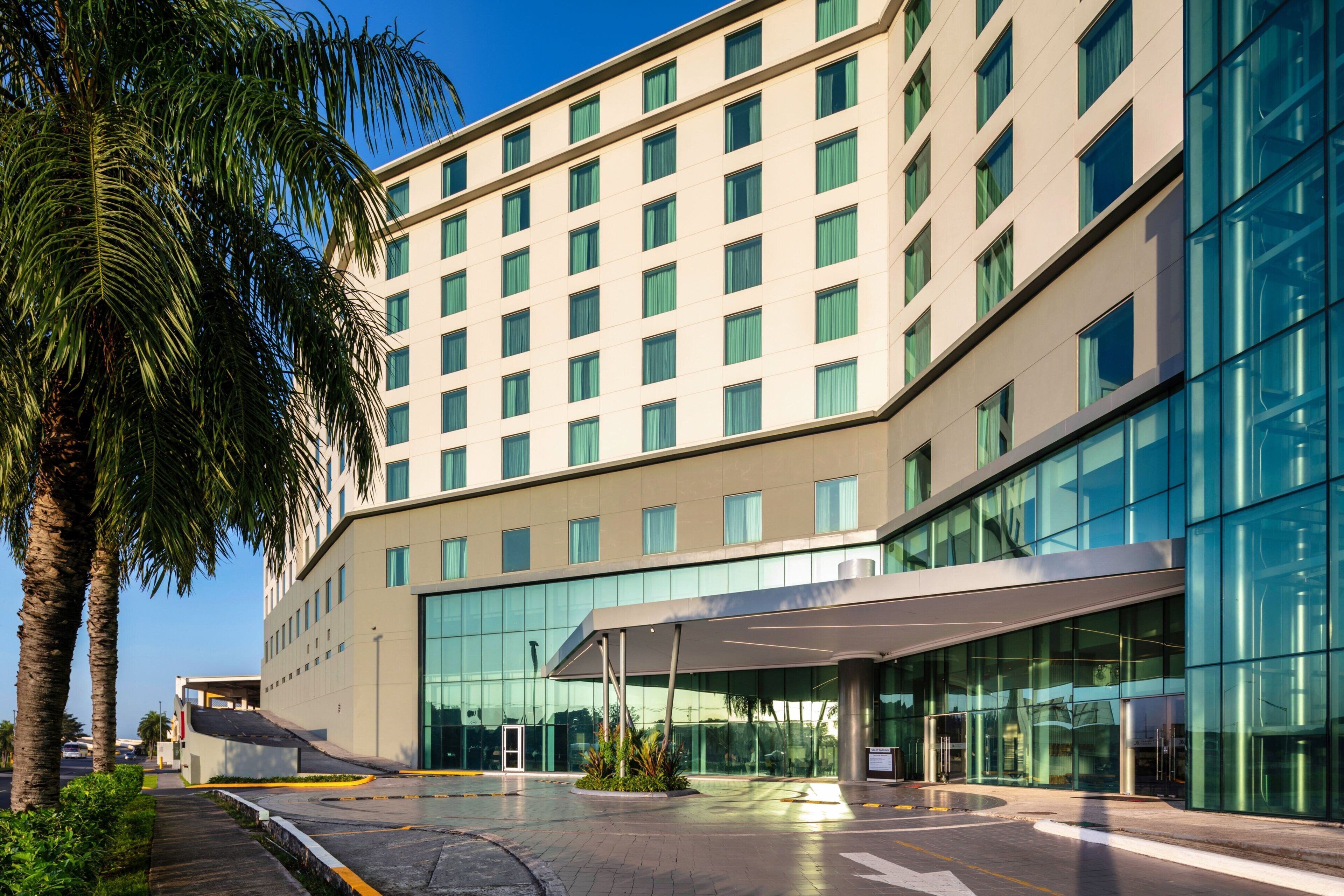 Un hotel moderno con fachada de vidrio, un acceso cubierto y palmeras en primer plano.
