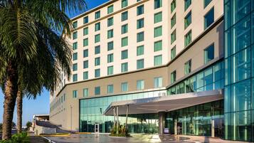 Un hotel moderno con fachada de vidrio, un acceso cubierto y palmeras en primer plano.