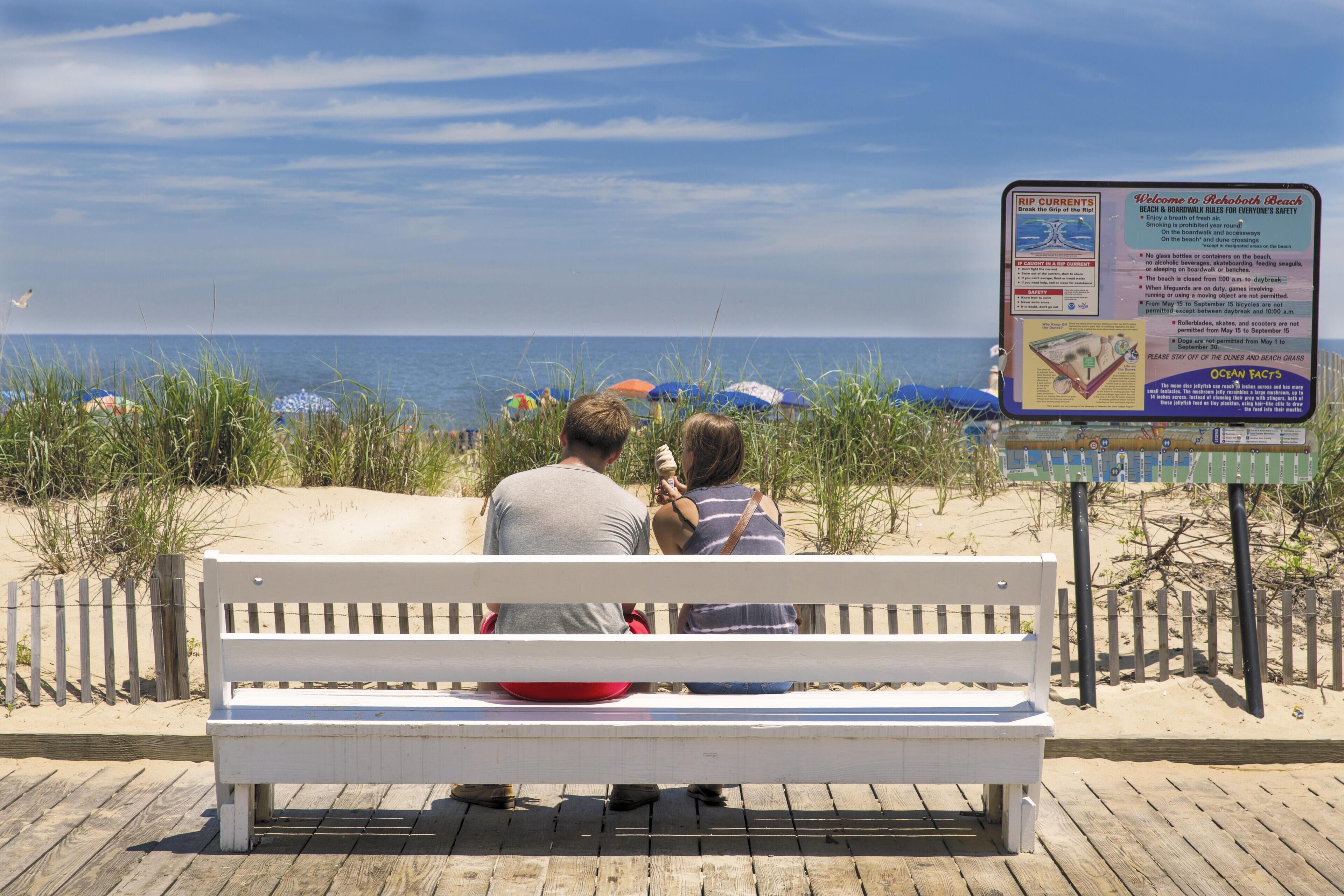 A wooden bench with a white backrest and a metal frame, positioned on a wooden deck with a sandy beach in the background.