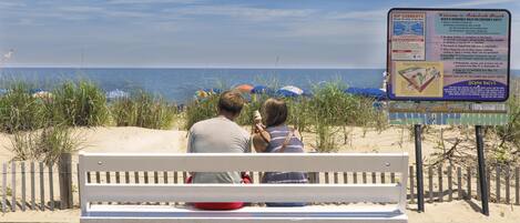 A wooden bench with a white backrest and a metal frame, positioned on a wooden deck with a sandy beach in the background.