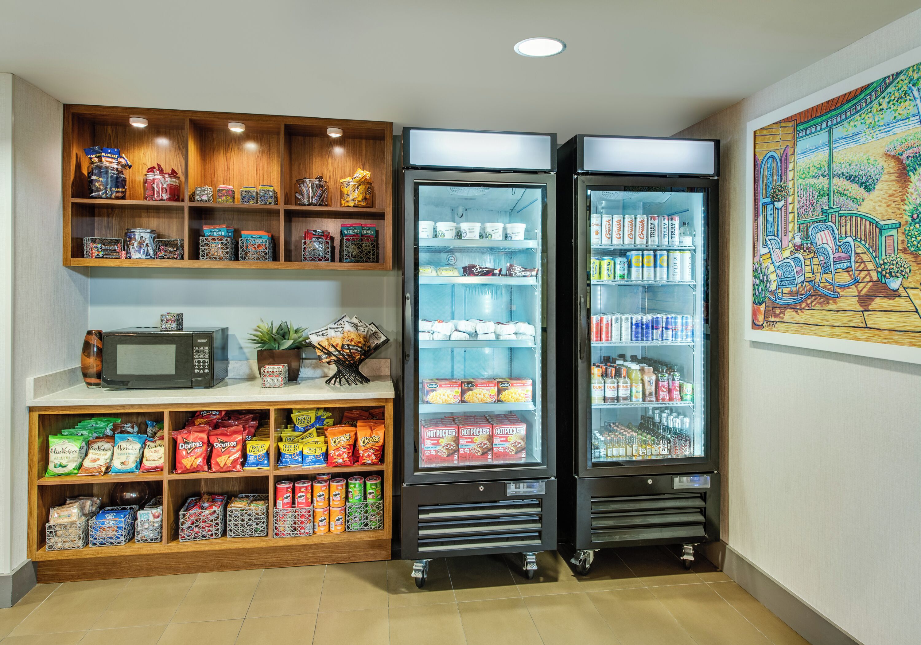 A kitchenette with a microwave, a refrigerator, and shelves stocked with snacks and beverages.