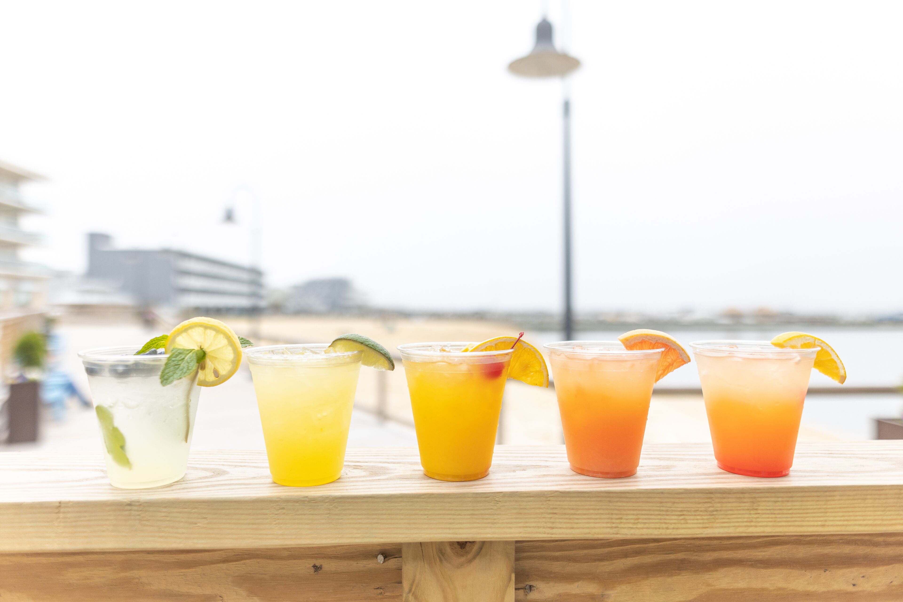 A wooden bar counter with five glasses of various colored beverages, each garnished with a slice of fruit and mint leaves.