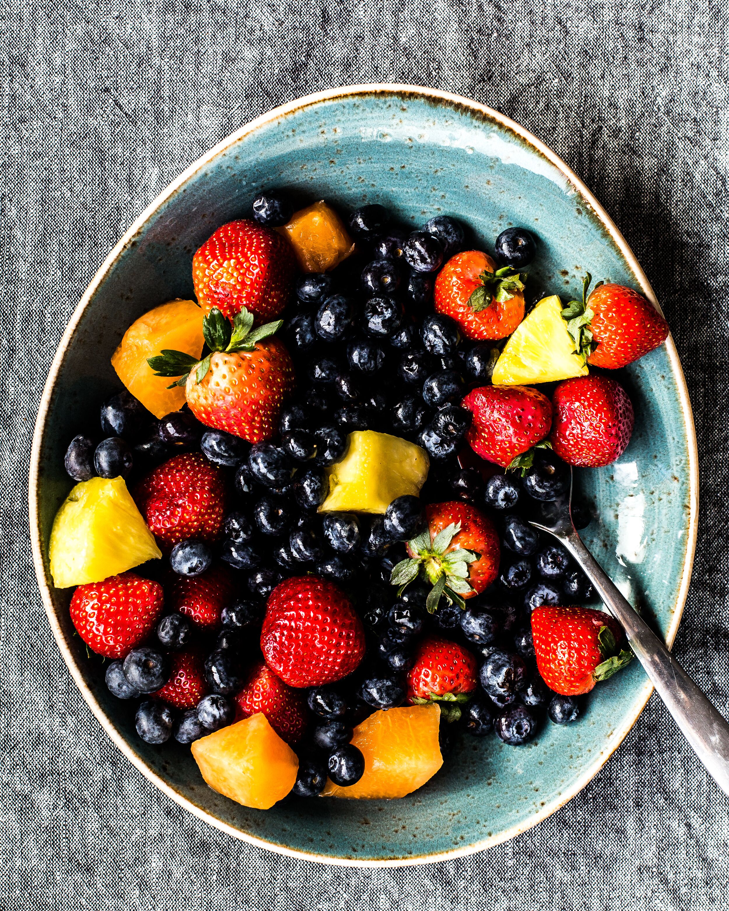 A bowl of mixed berries and pineapple chunks on a textured fabric surface.