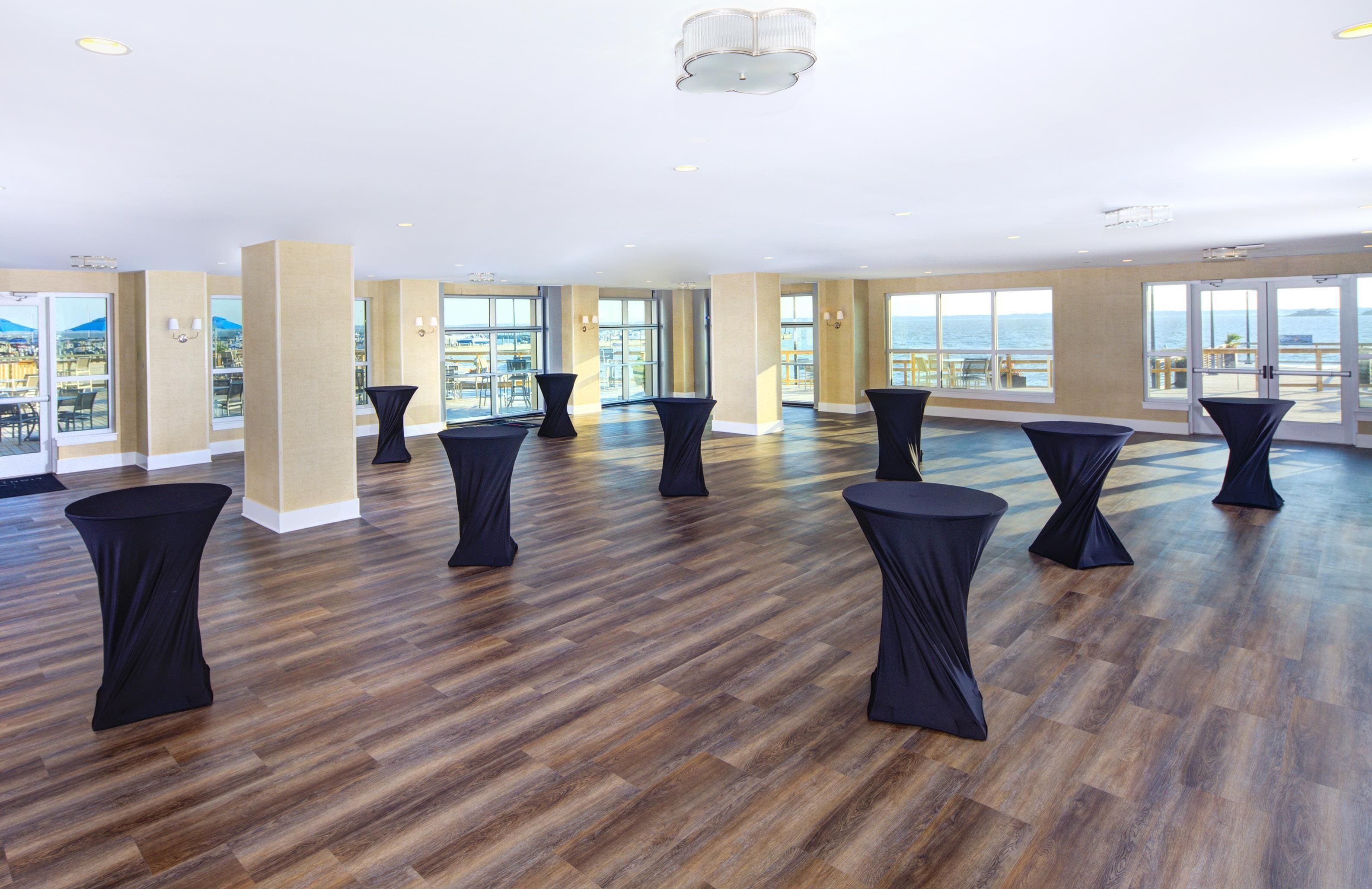 A spacious room with wooden flooring, large windows, and black-covered tables.
