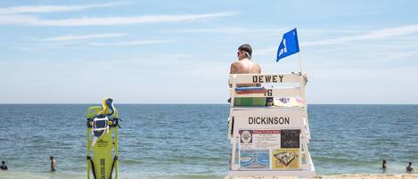 Una torre de socorrista con los nombres DEWEY y DICKINSON, una bandera azul y una tabla de surf verde apoyada en ella.