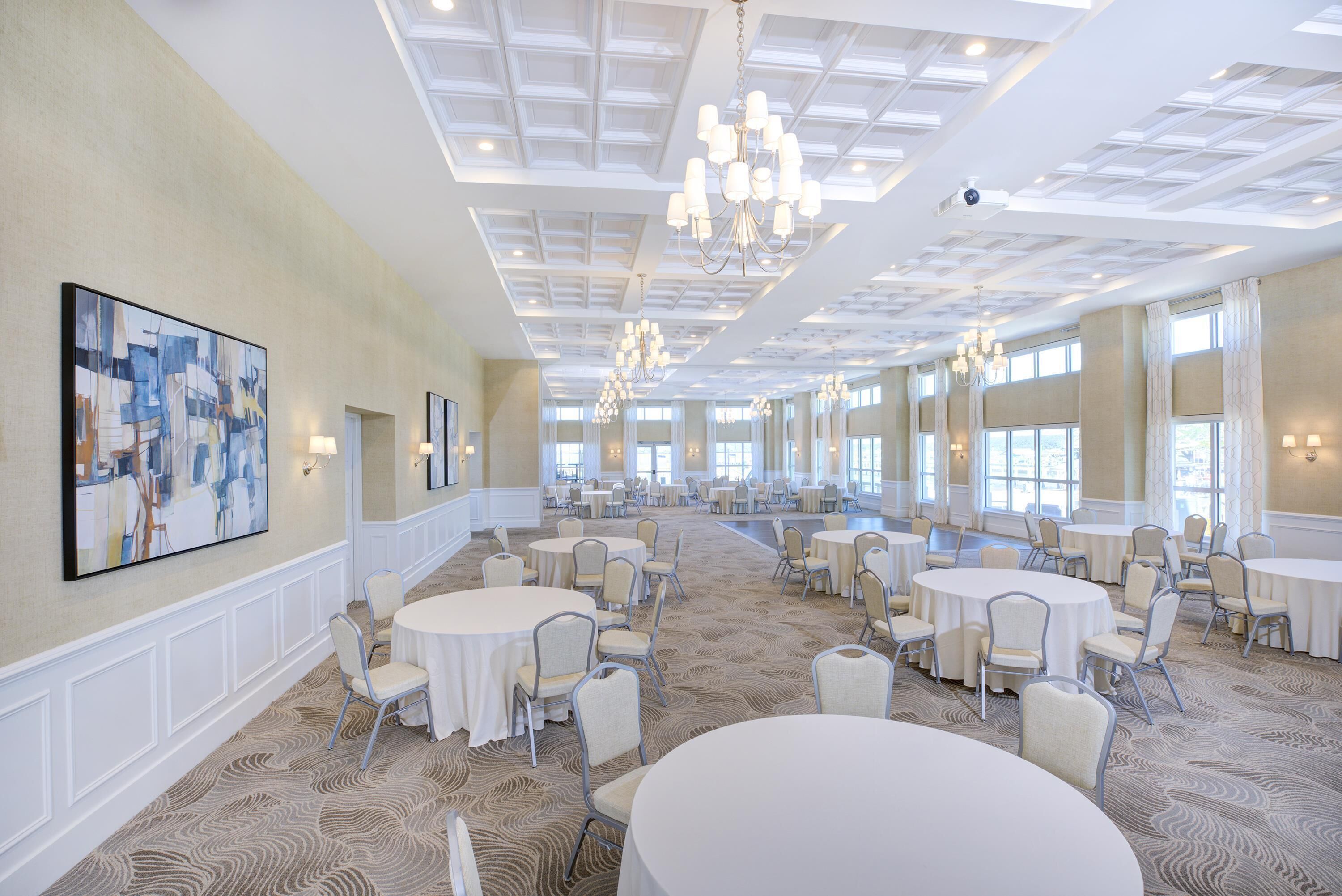 A spacious banquet hall with round tables and chairs, large windows, and a high ceiling with chandeliers.