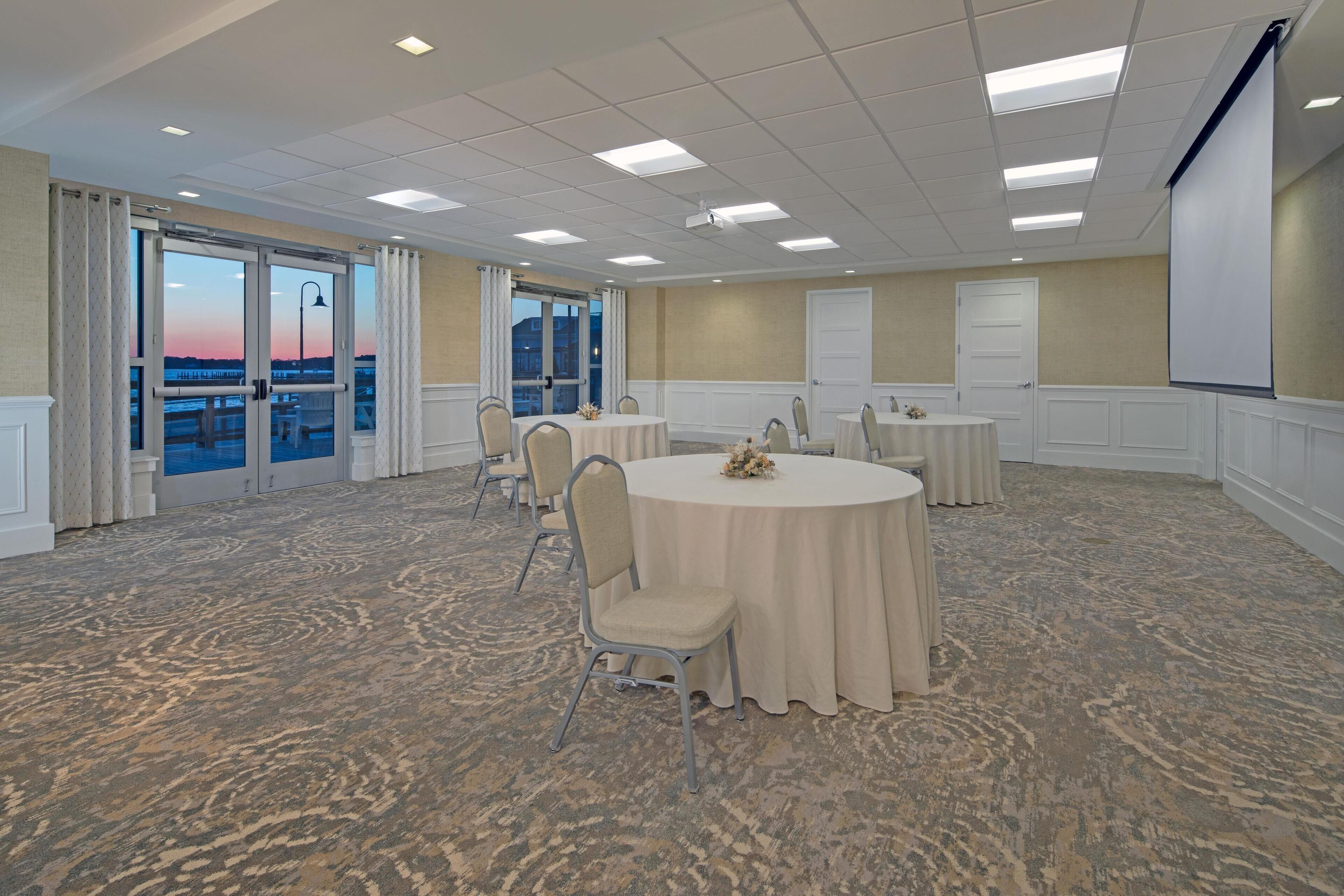 A conference room with a large table, chairs, a projector screen, and a view of the sunset through the glass doors.