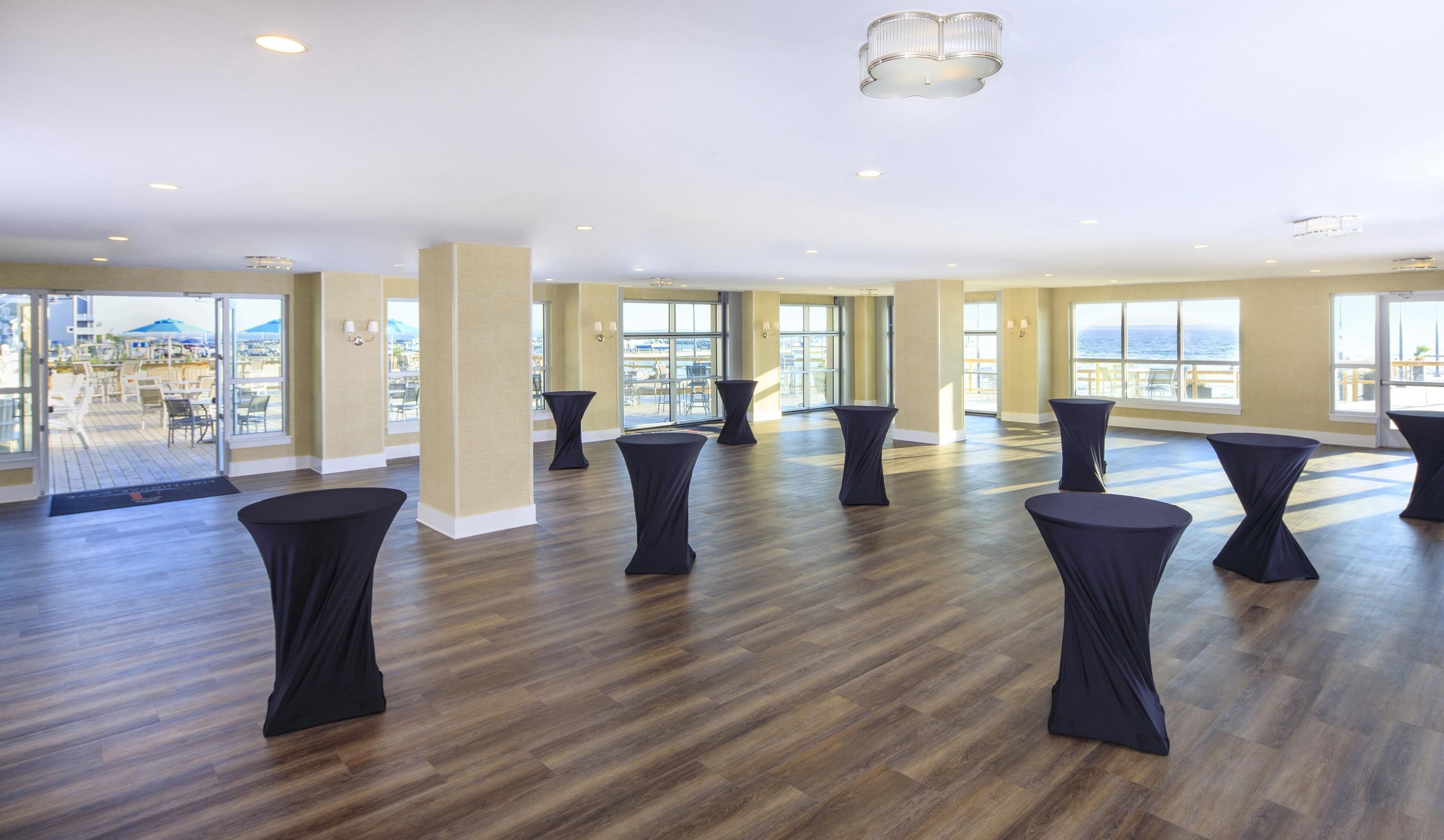 A spacious room with wooden flooring, multiple round tables, and large windows offering a view of the ocean.