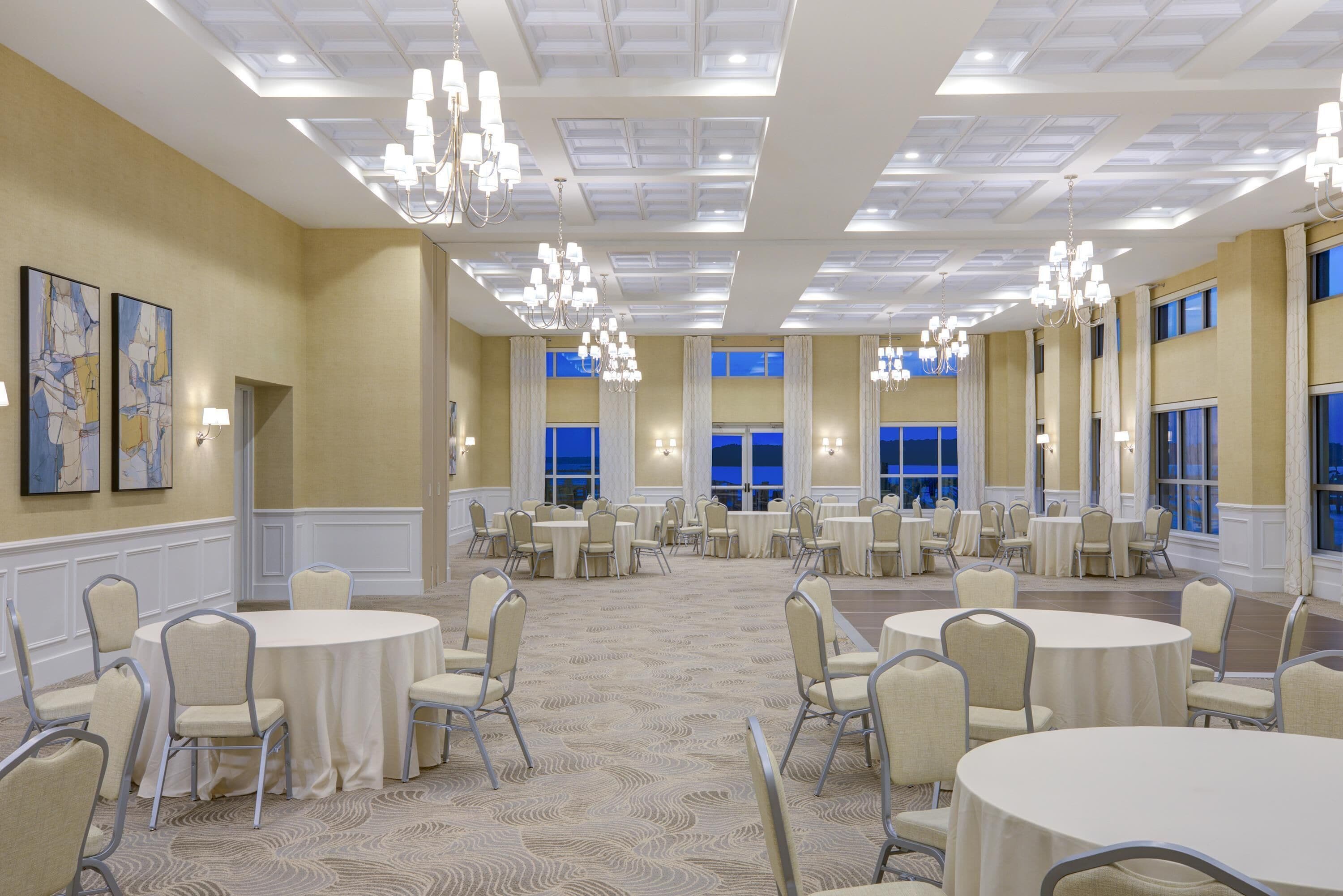 A large banquet hall with round tables and chairs, chandeliers, and large windows.