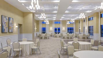 Un amplio salón de banquetes con mesas y sillas redondas, candelabros y grandes ventanales.