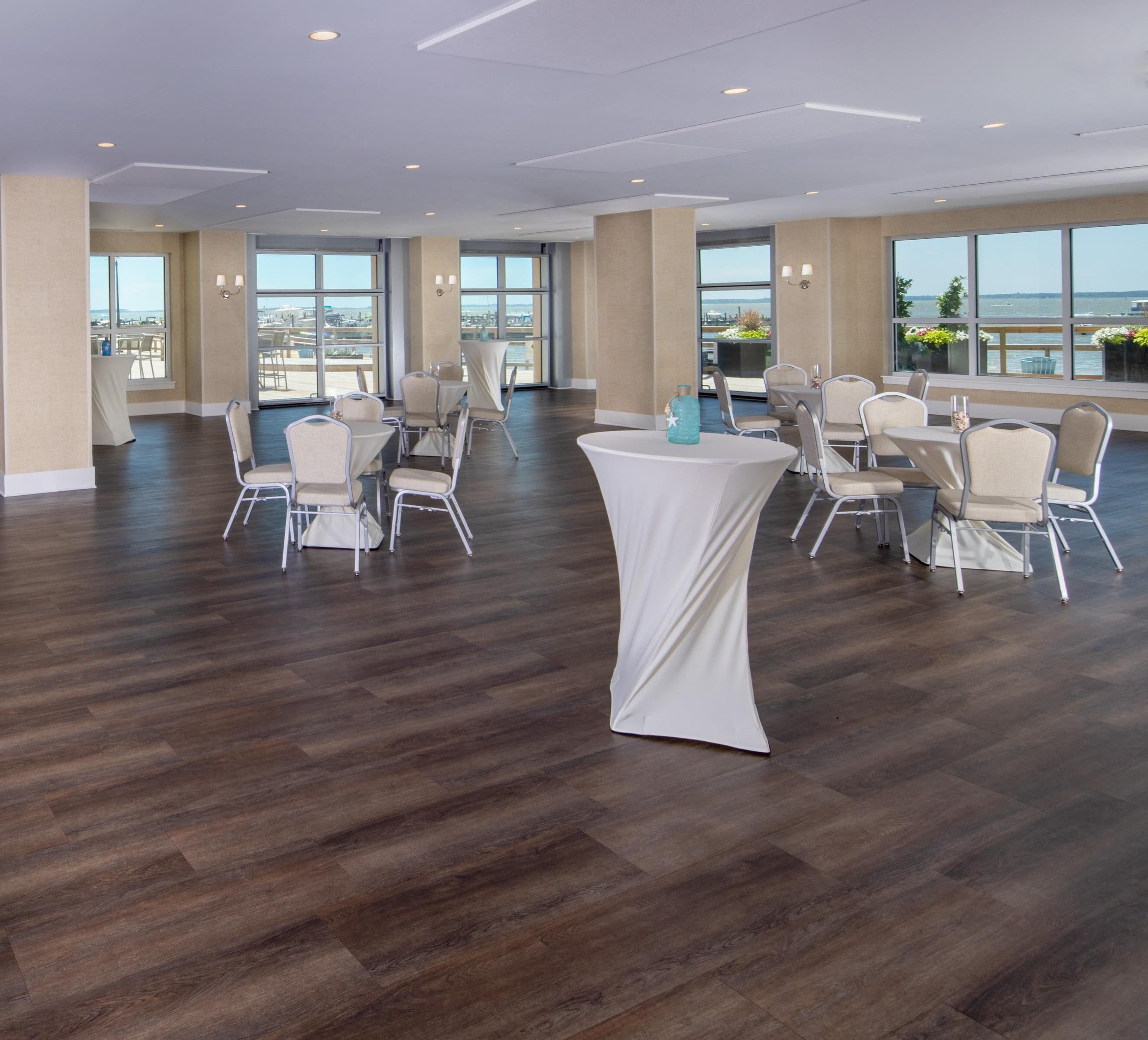 Un vaste espace intérieur doté de plusieurs tables et chaises rondes, de grandes fenêtres offrant une vue sur la ville et d’un plancher en bois.
