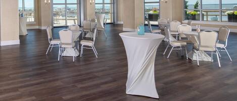 A spacious indoor area with multiple round tables and chairs, large windows offering views of the city, and a wooden floor.