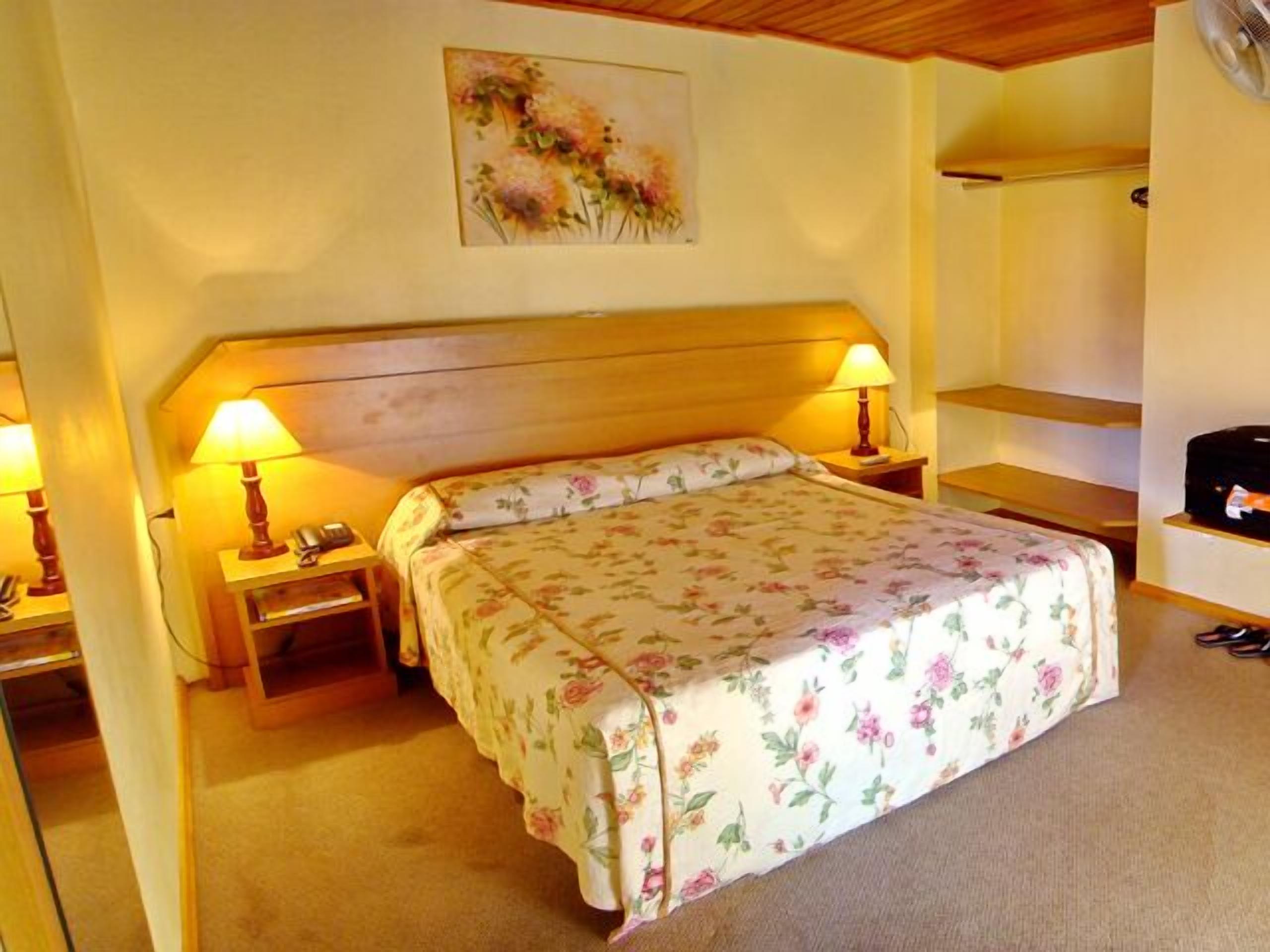 A bedroom with a wooden headboard, two bedside tables with lamps, a floral bedspread, and a painting on the wall.