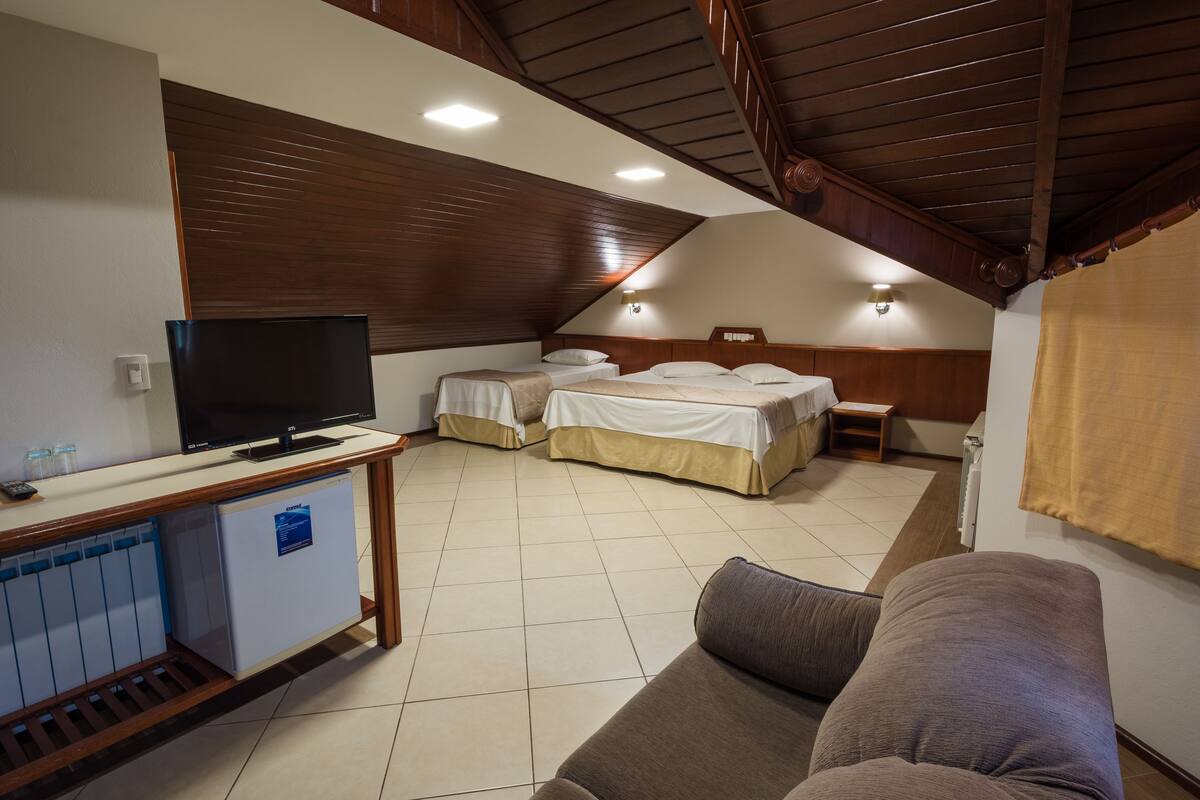 Ett hotellrum med TV, ett litet kylskåp, två sängar, en soffa och ett träpanelat tak.
