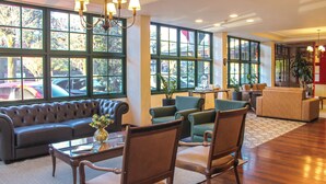 A spacious lobby with a leather sofa, wooden chairs, a coffee table, and large windows offering a view of parked cars outside. - Hotel Fioreze Origem (Gramado)