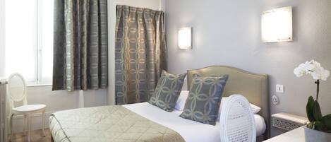 A hotel room with a bed, a chair, a desk, a potted plant, and a window with curtains.