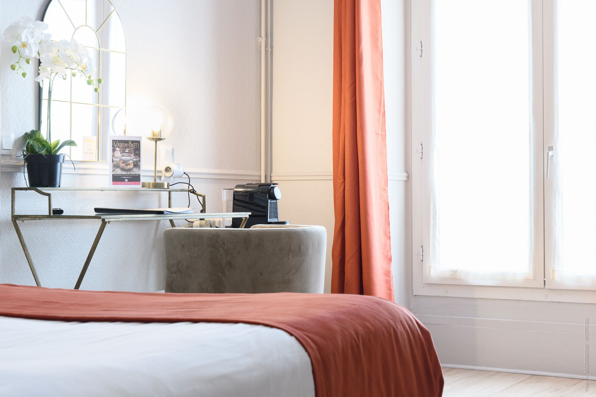 A hotel room with a bed, a desk with a lamp, a potted plant, and a window with curtains.