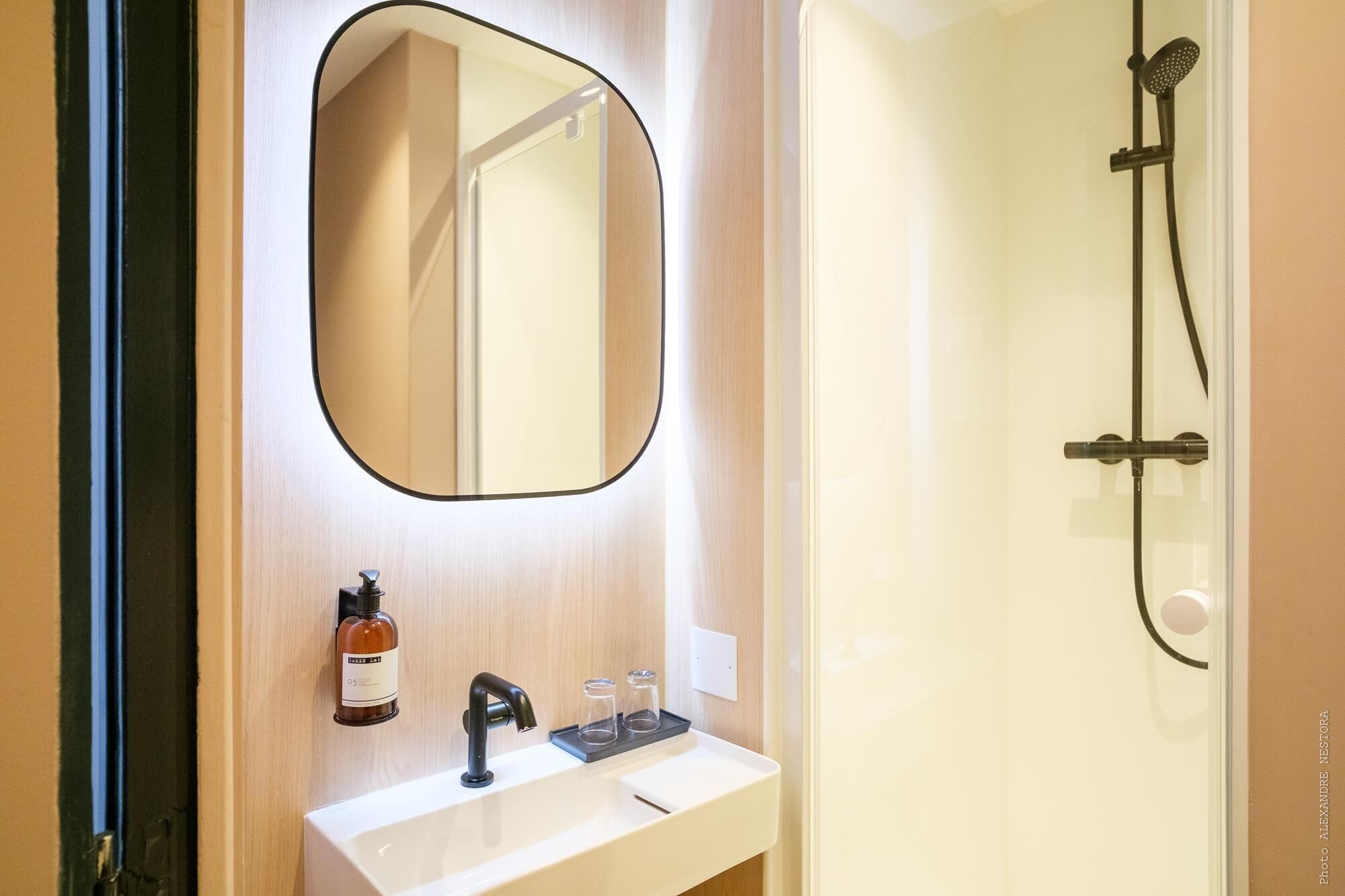A bathroom with a white sink, a mirror, a shower, and a bottle of soap.