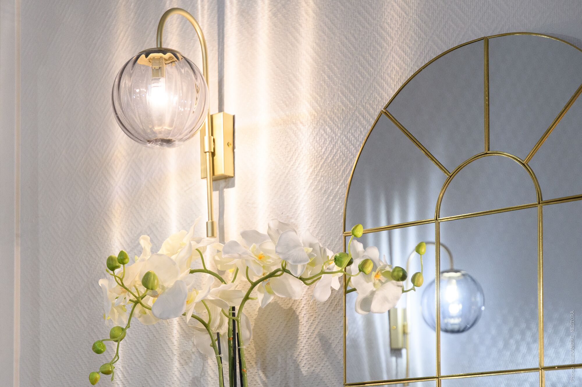 En väggmonterad lampa med en genomskinlig lampskärm, en spegel med guldfärgad ram och en vas med vita orkidéer.