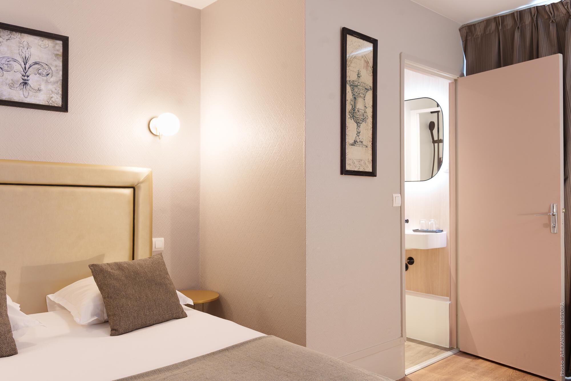 Habitación de hotel con una cama, una mesita de noche, una lámpara en la pared y una puerta que da al baño.