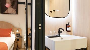 A hotel room with a bed, bedside table, lamp, and a bathroom with a sink, mirror, and toiletries.