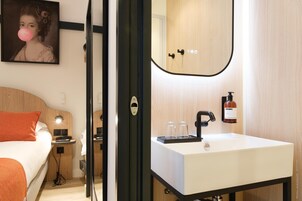 A hotel room with a bed, bedside table, lamp, and a bathroom with a sink, mirror, and toiletries.