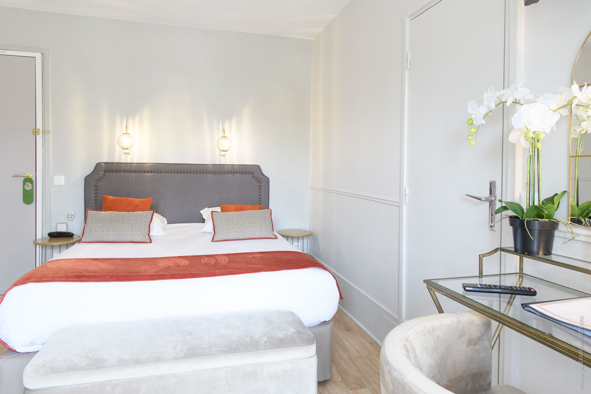 A hotel room with a large bed, two bedside tables, a glass desk with a chair, a mirror, and a potted plant.