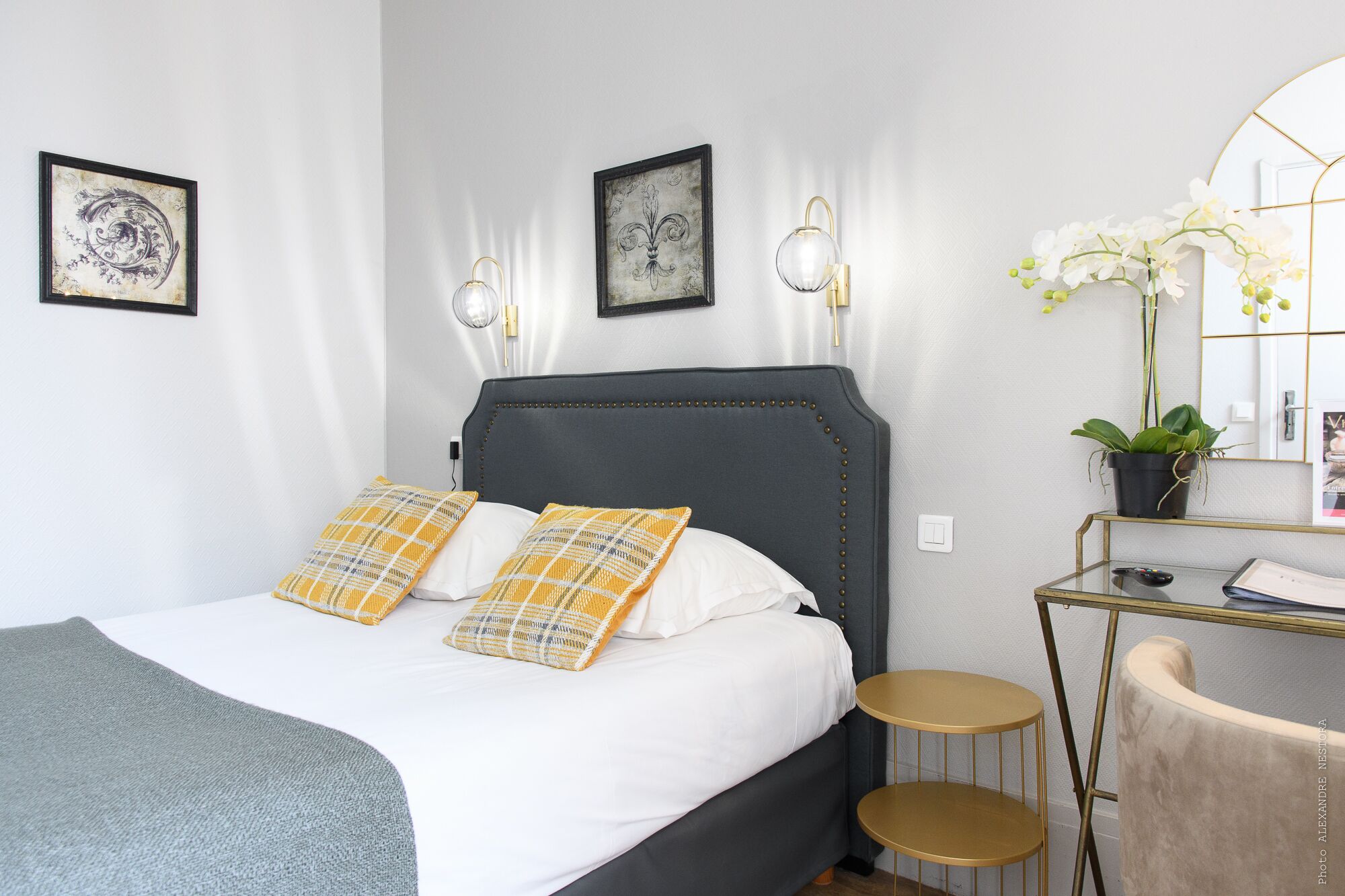 Ein ordentlich eingerichtetes Schlafzimmer mit einem Bett, zwei Wandleuchten, einem Nachttisch mit Pflanze und einem Spiegel.
