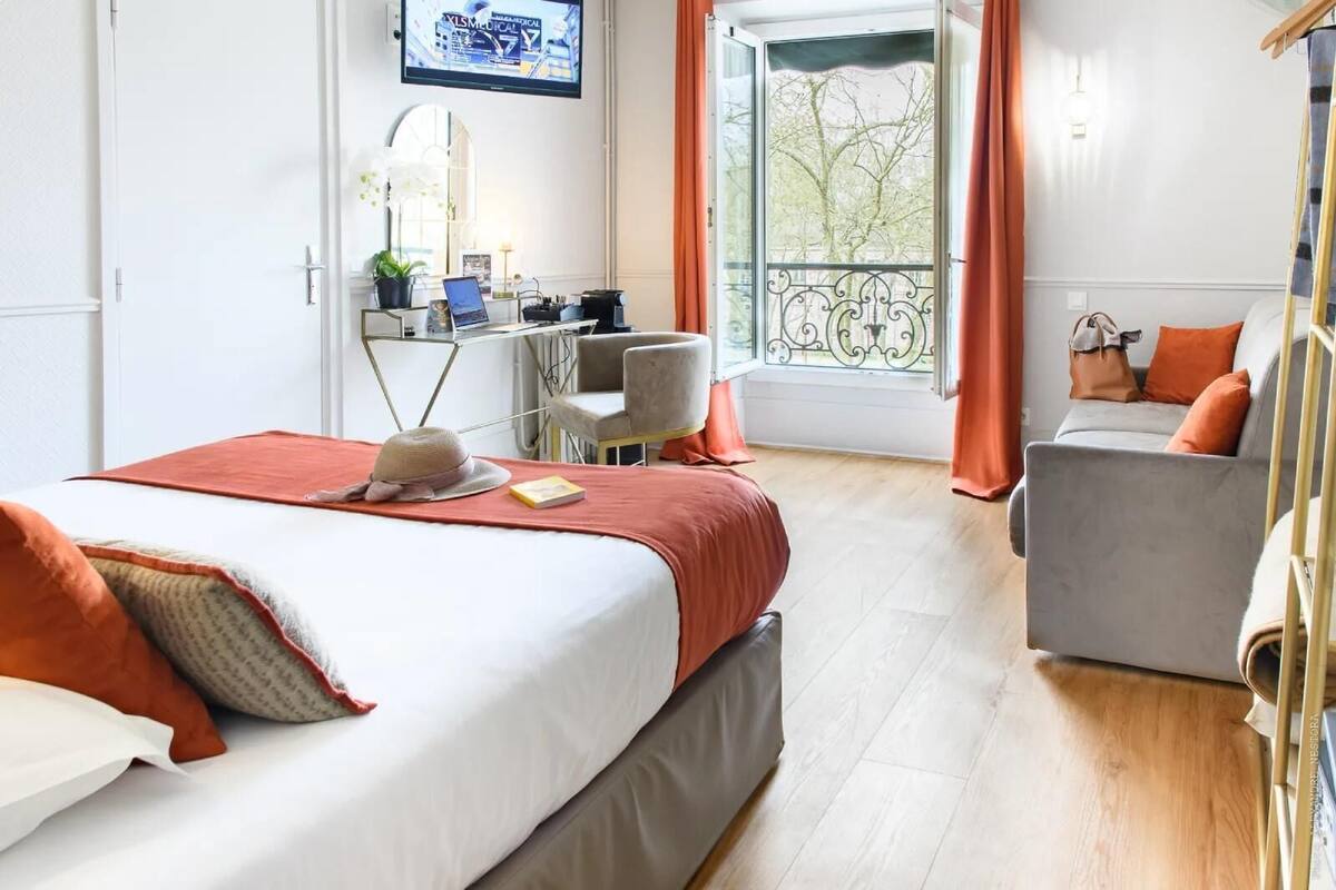 Quarto de hotel com cama, escrivaninha, cadeira e varanda com cortinas.