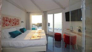 A modern hotel room with a large bed, a glass shower, a red chair, and a view of the sea.