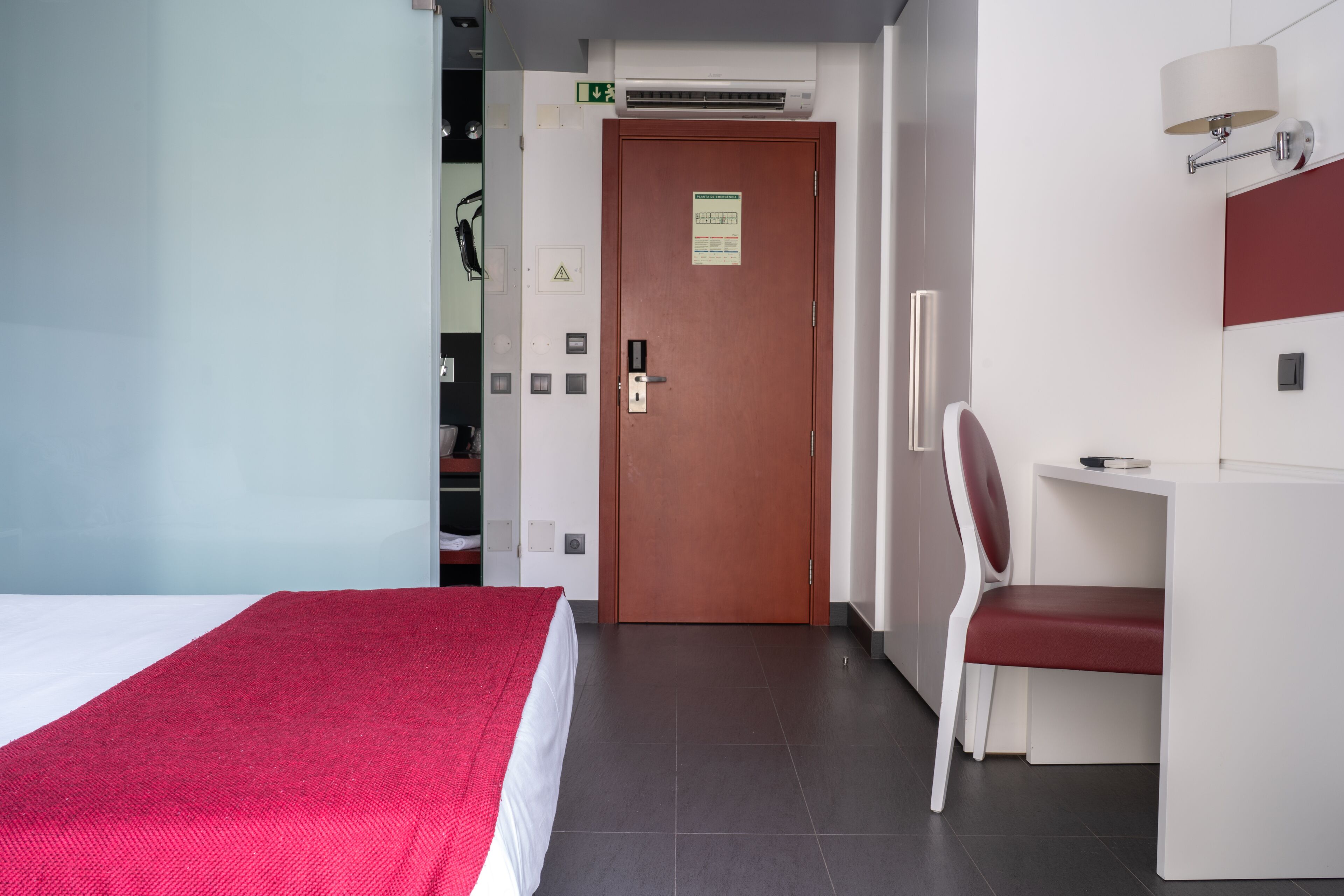 A hotel room with a bed, a desk, a chair, a lamp, and a door with an emergency exit sign.