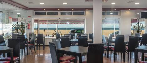 A restaurant with tables and chairs arranged for dining, a view of the beach through large windows, and a fire extinguisher mounted on the wall.