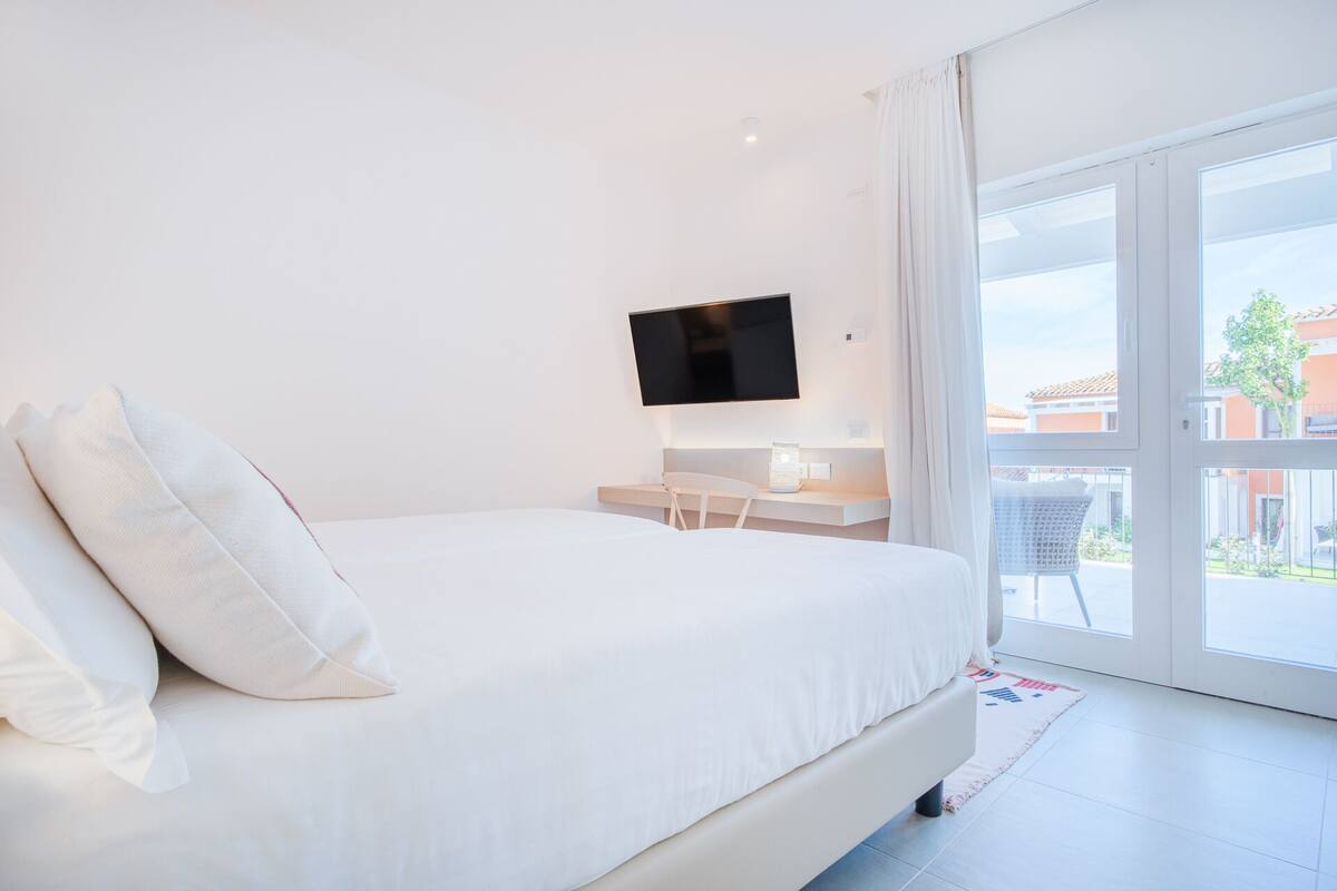 Un dormitorio con cama, escritorio, televisor y balcón con vistas a edificios.