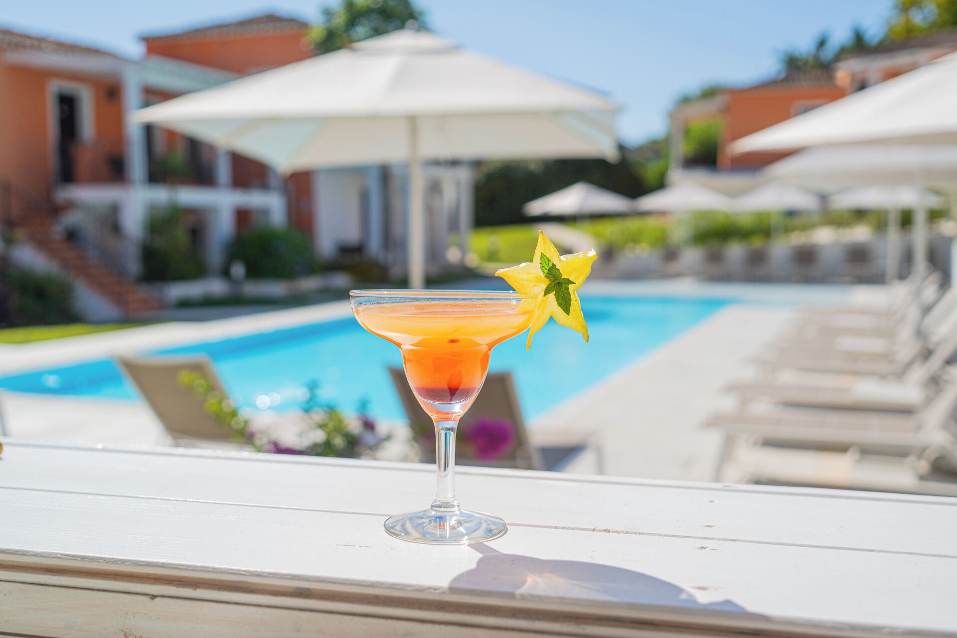 Ein Cocktail auf einem weißen Tisch in der Nähe eines Pools mit Sonnenschirmen und Liegestühlen.