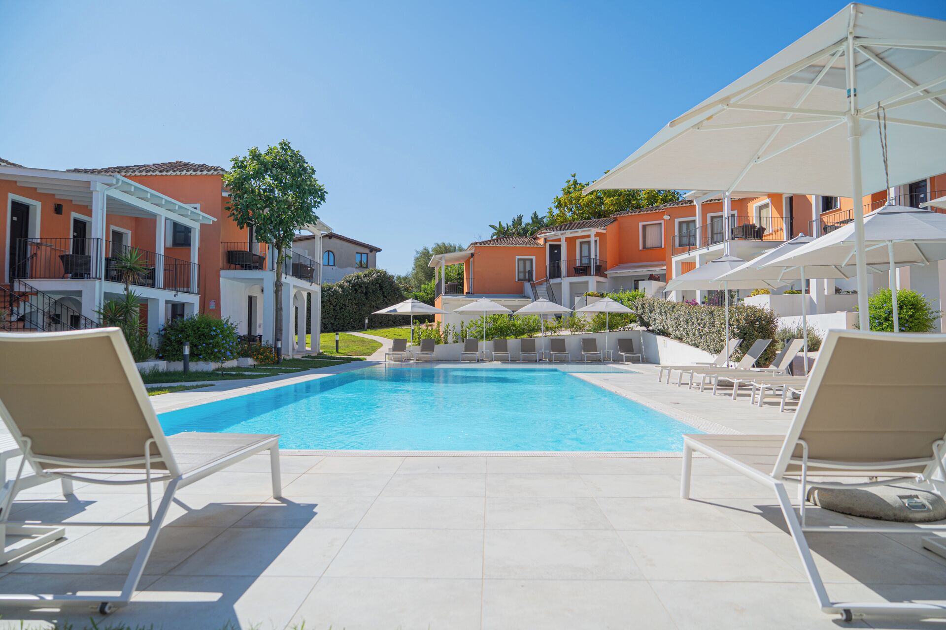 Una piscina con lettini e ombrelloni, circondata da edifici con balconi e un cielo sereno.