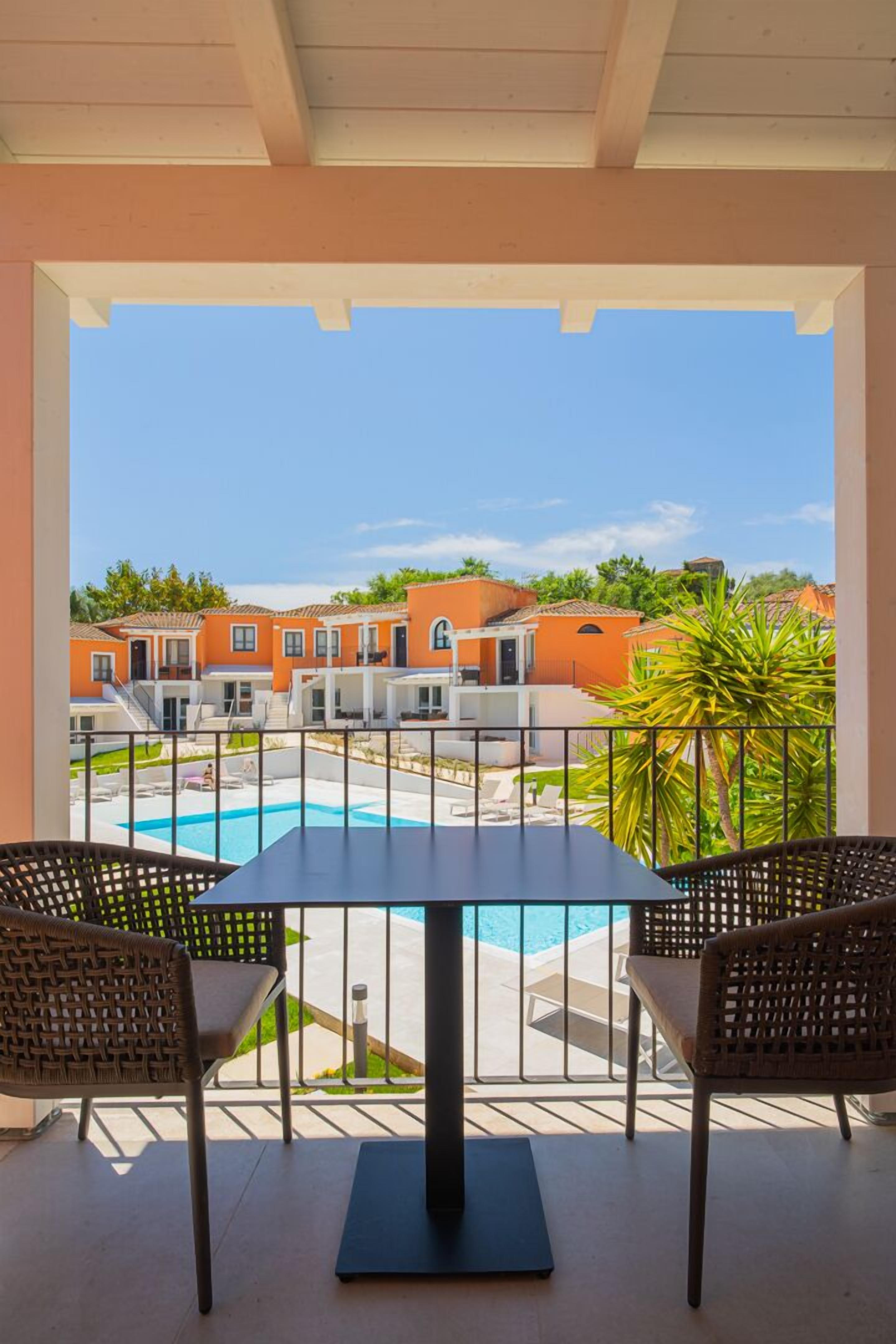A balcony with two wicker chairs, a table, and a view of a swimming pool and orange buildings in the background.