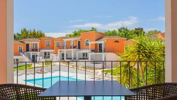 A balcony with two wicker chairs, a table, and a view of a swimming pool and orange buildings in the background.