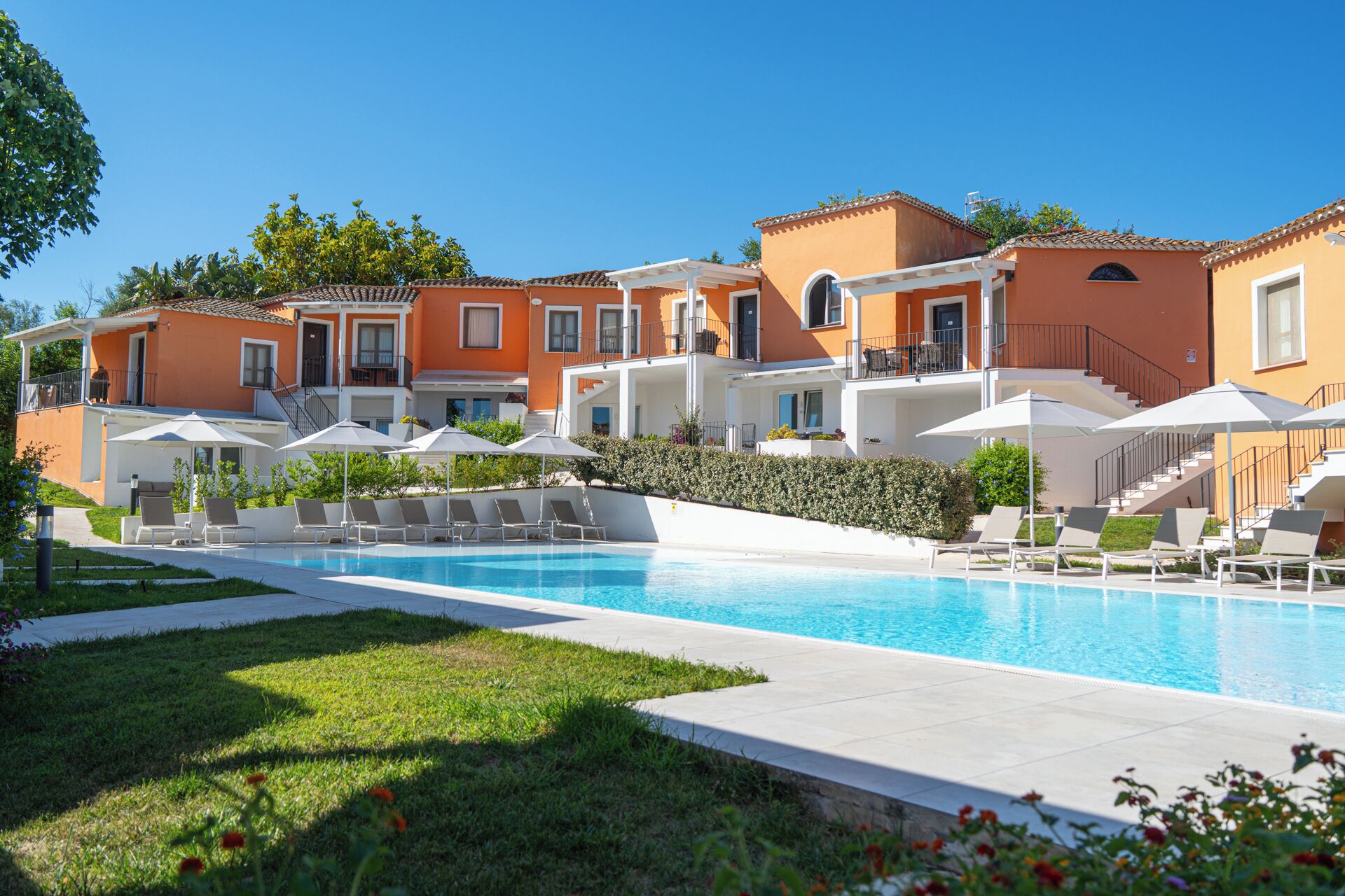 Un espace piscine doté de chaises longues et de parasols, entouré de bâtiments aux façades orange et dotés de balcons.