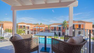 A covered outdoor area with wicker chairs and a table, overlooking a swimming pool and orange buildings.