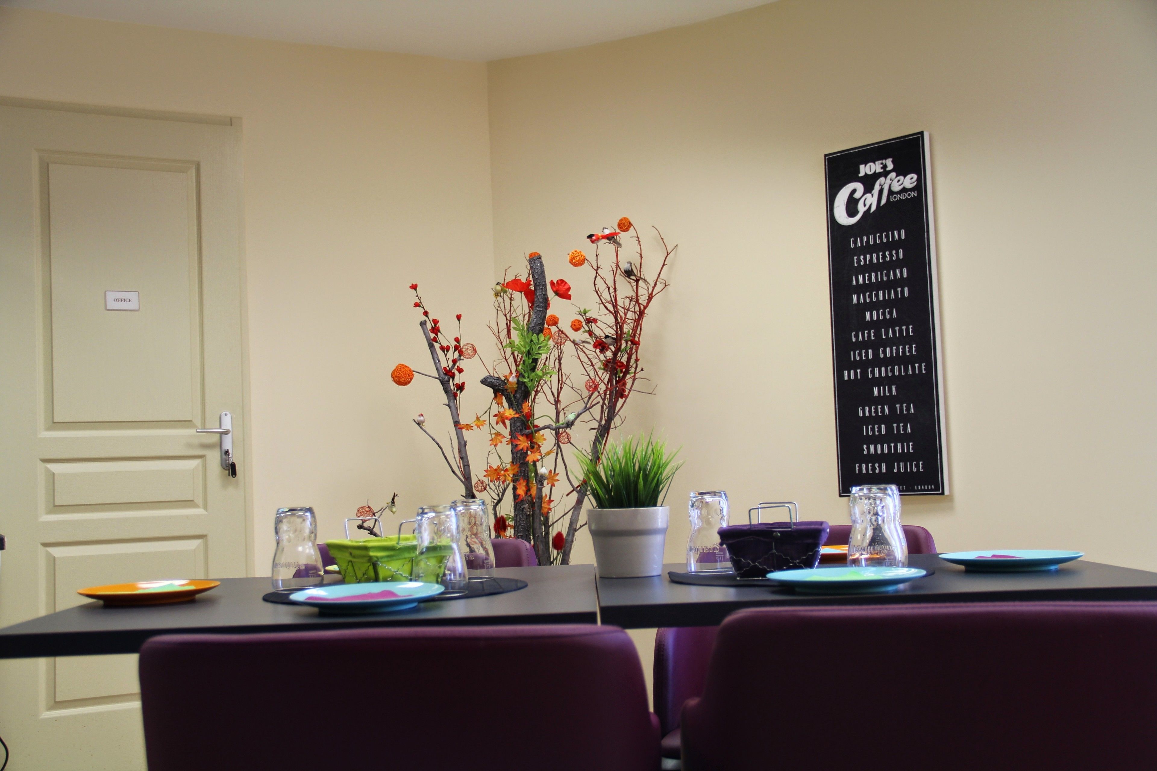 Un coin repas avec une table dressée pour deux personnes, une plante en pot et un panneau affichant les différentes sortes de café proposées.