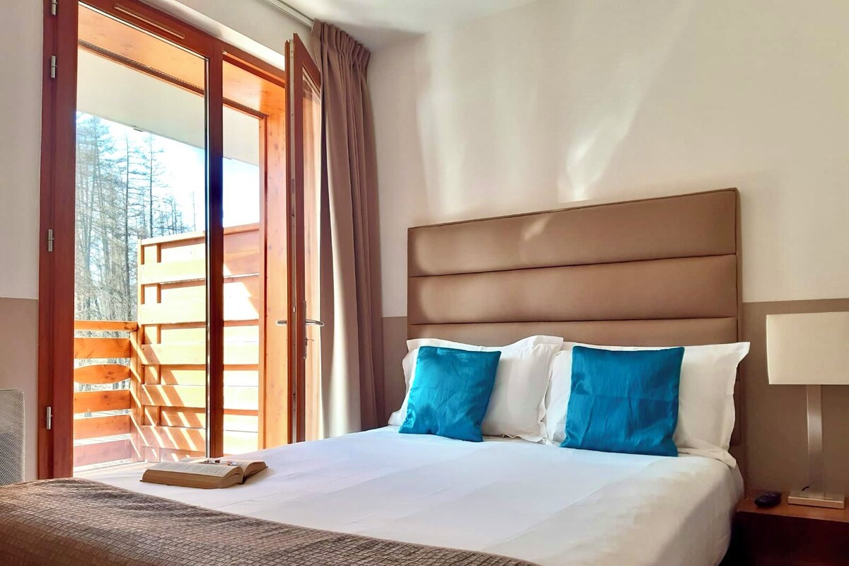 Habitación de hotel con una cama grande, cabecera de madera y vistas a una terraza de madera y árboles a través de una puerta corrediza de vidrio.