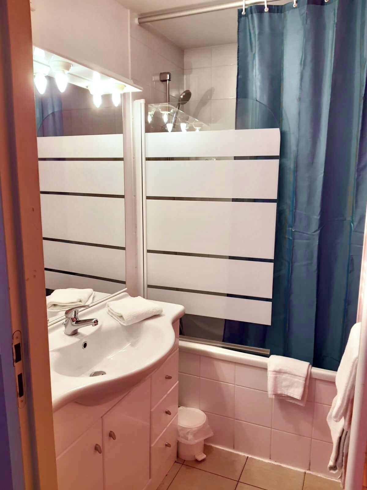 Un baño con lavamanos, espejo y tocador blancos, una ducha con cortina azul y un inodoro blanco.