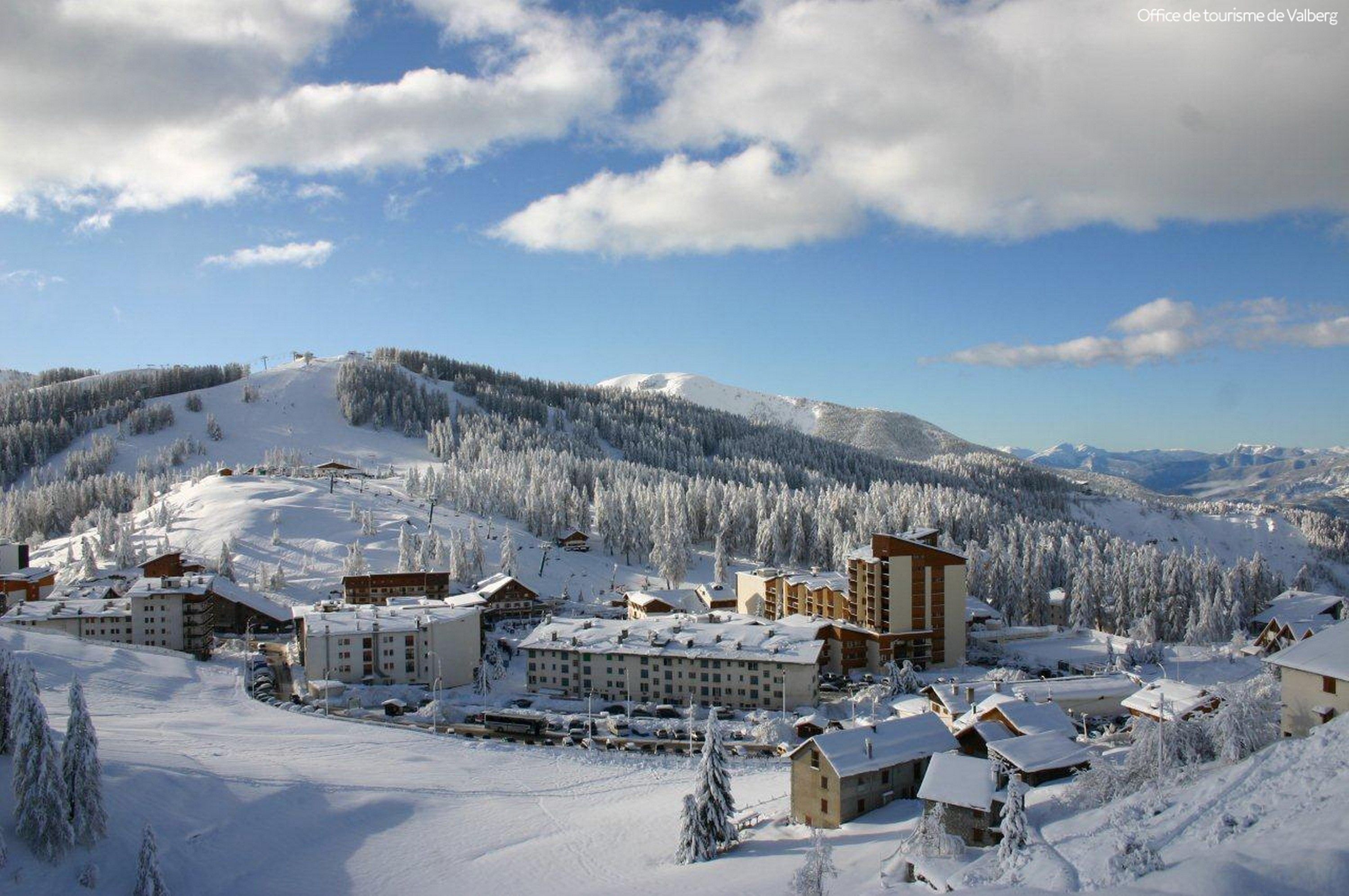 A snow-covered resort with multiple buildings, including a large lodge and smaller cabins, set against a backdrop of snow-laden mountains and a clear sky.