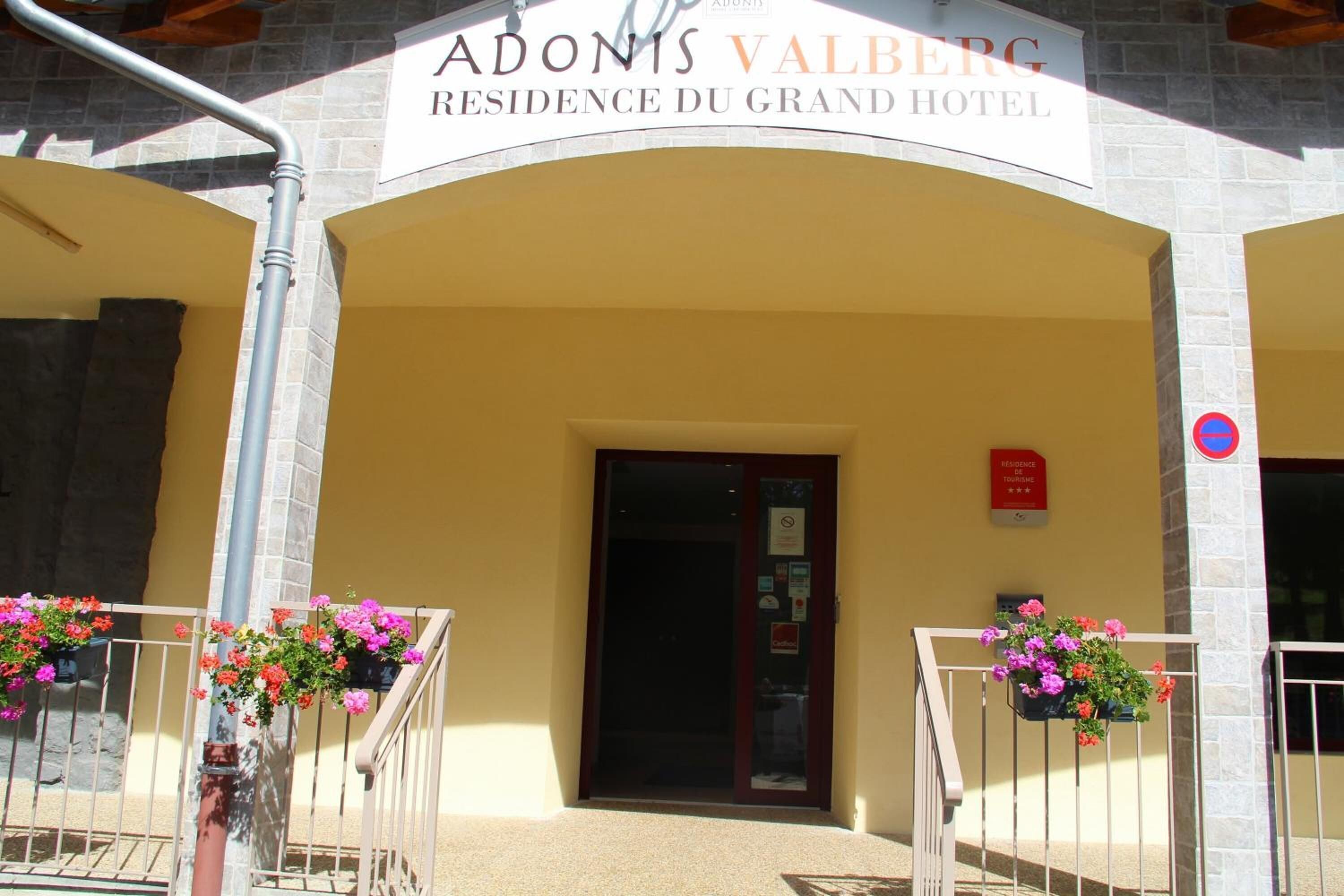 Das äußere Erscheinungsbild des Adonis Valberg Residence du Grand Hotel mit einer Steinfassade, einer dunklen Eingangstür und Blumenkörben an den Geländern.