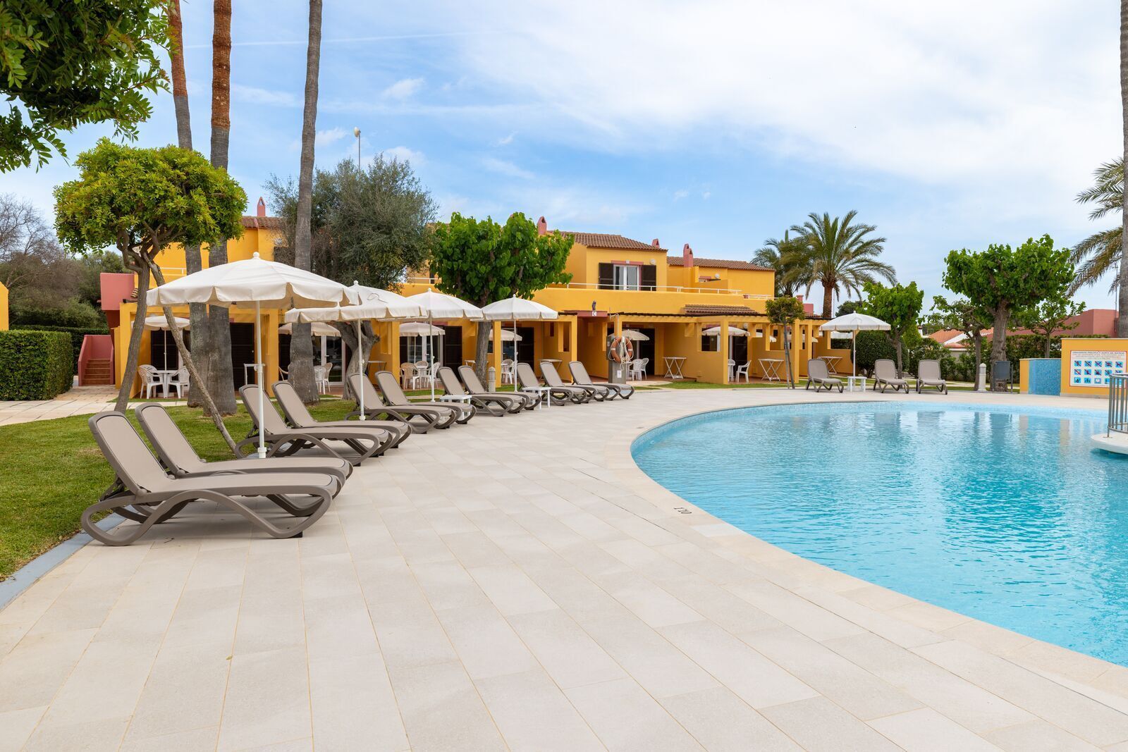 A poolside area with lounge chairs and umbrellas, a clear blue pool, and a yellow building in the background.